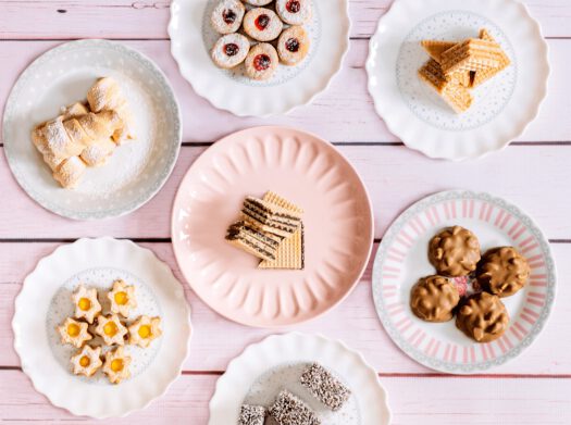 Siedem rodzajów ciastek z oferty Cukierni Urbańscy, przedstawionych na osobnych talerzykach w pastelowych kolorach, na różowych deskach.