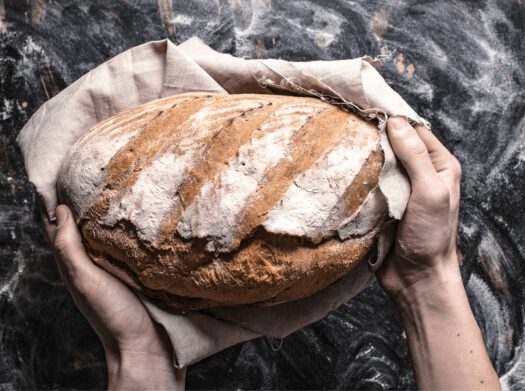 Duży bochenek chleba w ściereczce trzymanej przez mężczyznę - zdjęcie na tle stołu opruszonego mąką
