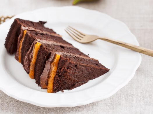 Kawałek tortu czekoladowego na zamówienie przekładanego dwoma rodzajami nadzienia, zaprezentowany na białym talerzyku ze złotym widelcem
