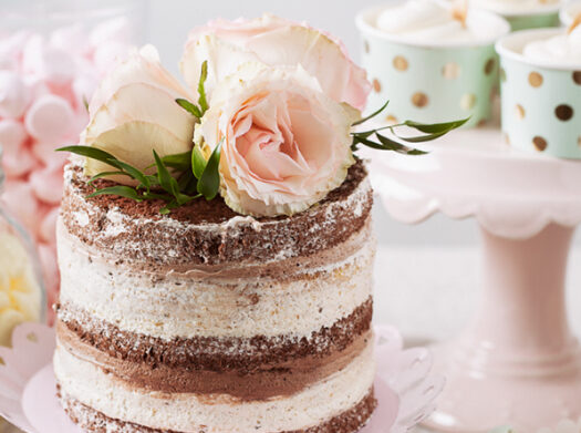 Tort nawiązujący do przyjęcia weselnego, ozdobiony rózowym kwiatem na górze oraz z elementami słodkiego stołu w tle.